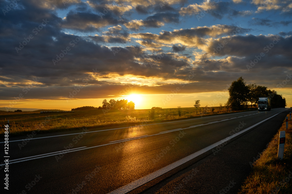 Sunset over the asphalt road in a rural landscape, a white truck coming from afar.