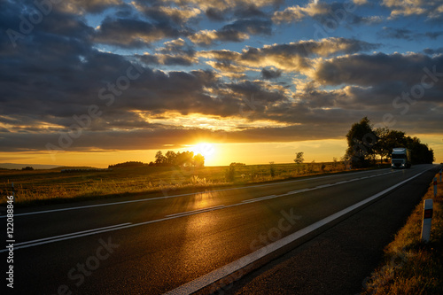 Obraz na płótnie Sunset over the asphalt road in a rural landscape, a white truck coming from afar. w salonie