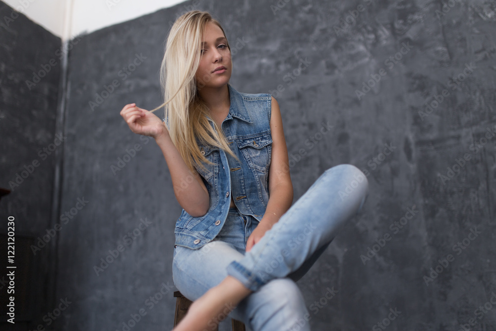 Portrait of the blonde in jeans and denim sleeveless jacket with her hair and natural makeup sitting on a wooden chair Portrait of the blonde in jeans and denim sleeveless jacket with her hair and natural makeup sitting on a wooden chair