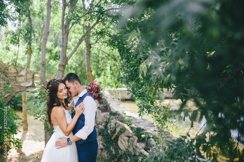 happy groom in a dark blue suit hugs a beautiful bride in a white wedding dress with pink and purple bouquet of flowers outdoors on the background of a stone arch happy groom in a dark blue suit hugs a beautiful bride in a white wedding dress with pink and purple bouquet of flowers outdoors on the background of a stone arch