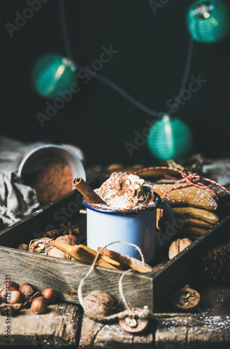 Mug of hot chocolate with whipped cream, cocoa powder, cinnamon, gingerbread cookies, nuts in wooden tray, dark wall with Christmas decoration garland of blue balls at background, selective focus photo