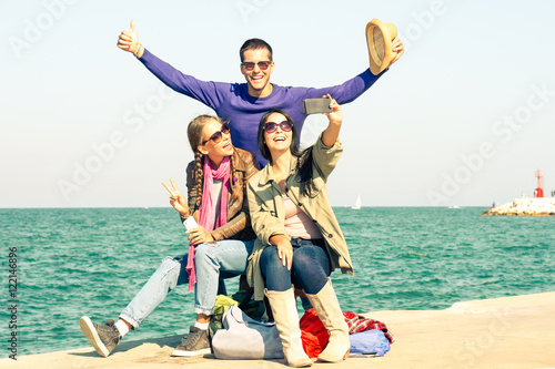 Cheerful friends having fun taking selfie with mobile at pier - Happy students smiling at camera for holiday photo on ocean background - Concept of teenager joyful moment outdoor