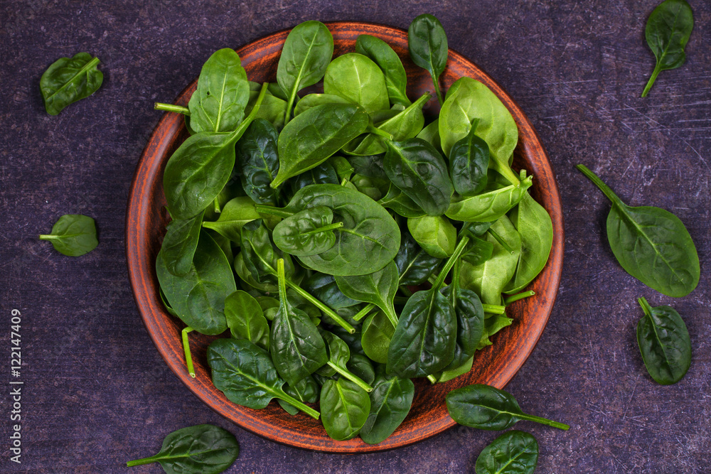 Green fresh baby spinach in rustic plate on dark wooden background. View from above, top studio shot Green fresh baby spinach in rustic plate on dark wooden background. View from above, top studio shot