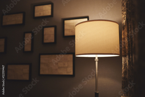A lamp in a dark corner of a hotel room with abstract frames out of focus on the wall behind it and a curtain to the right.