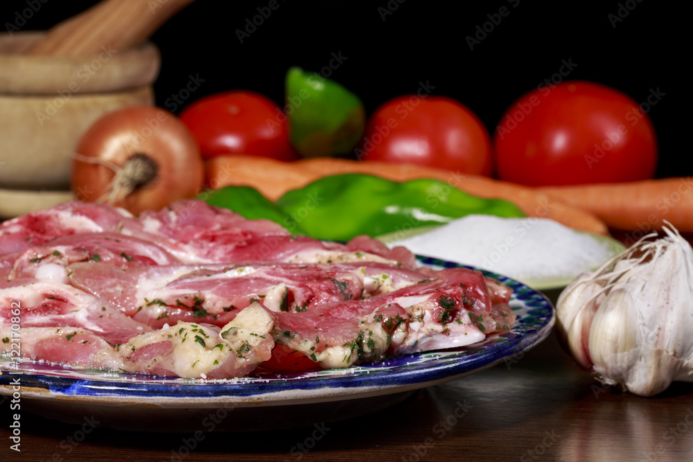 Dish with raw turkey meat marinated with garlic and parsley, beside garlic bulb, small dish with salt, green peppers and other vegetables. On black background