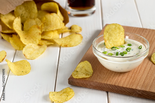 Potato chips in a paper bag and dipping sauce on a wooden table.