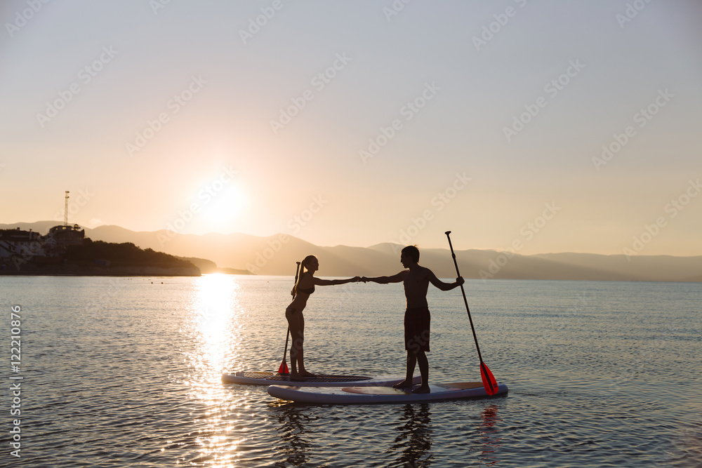 Fototapeta premium Silhouette of male and female on sup surf pull hands together at the ocean. Concept lifestyle, sport, love