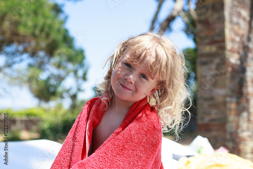 Adorable kid with curly blonde hair red towel sitting after swimming in outdoor pool 
