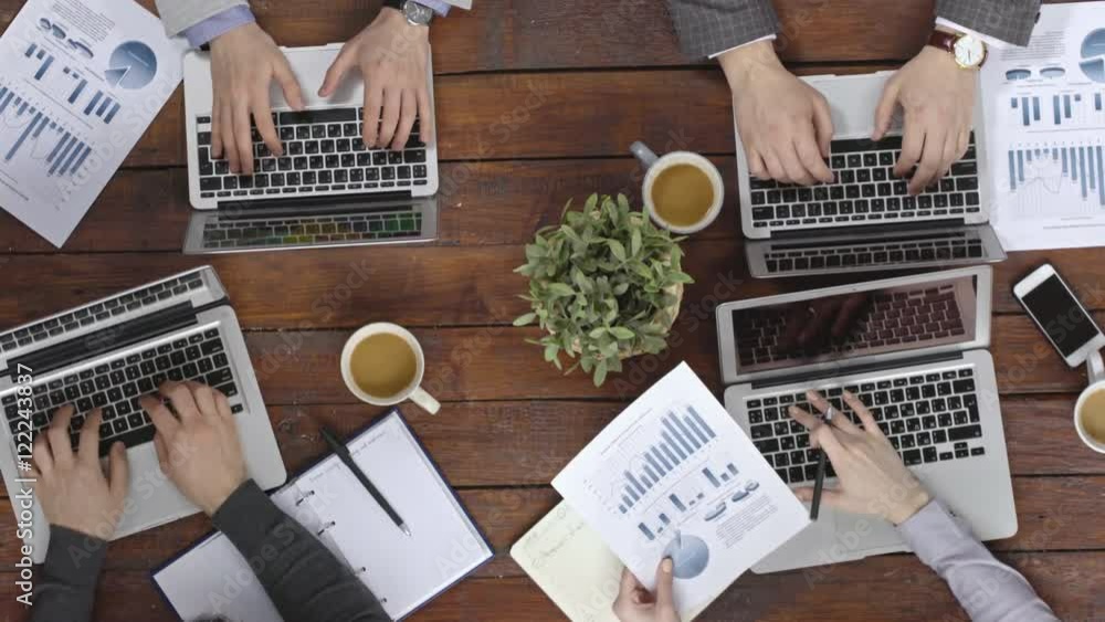 View from the top of hands of coworkers sitting at wooden table, typing on laptops and holding financial documents View from the top of hands of coworkers sitting at wooden table, typing on laptops and holding financial documents