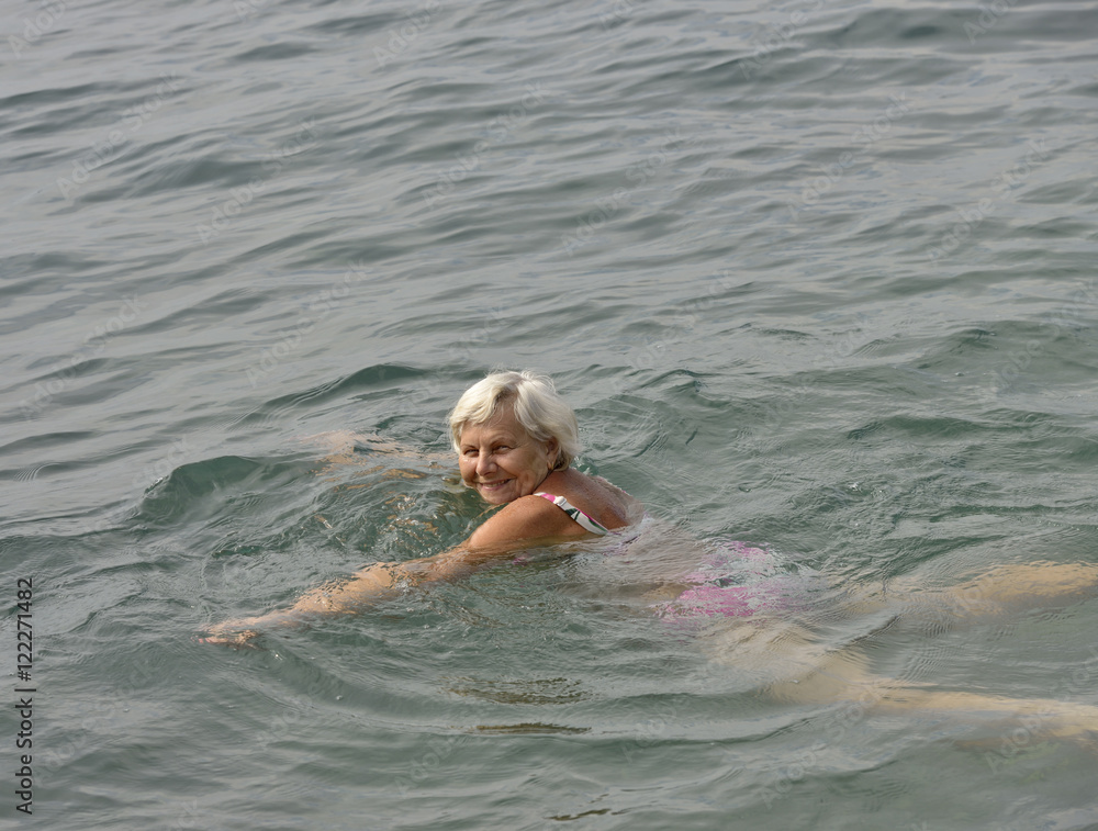 Aged smiling woman is swimming in light clear sea water.
