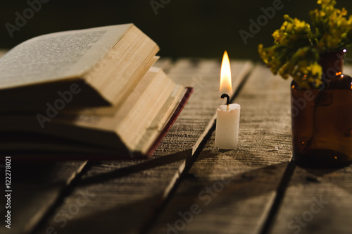 Thick book lying open on wooden surface, wax candle and small bottle with yellow flowers sitting next to it, beautiful night light setting, magic concept shoot