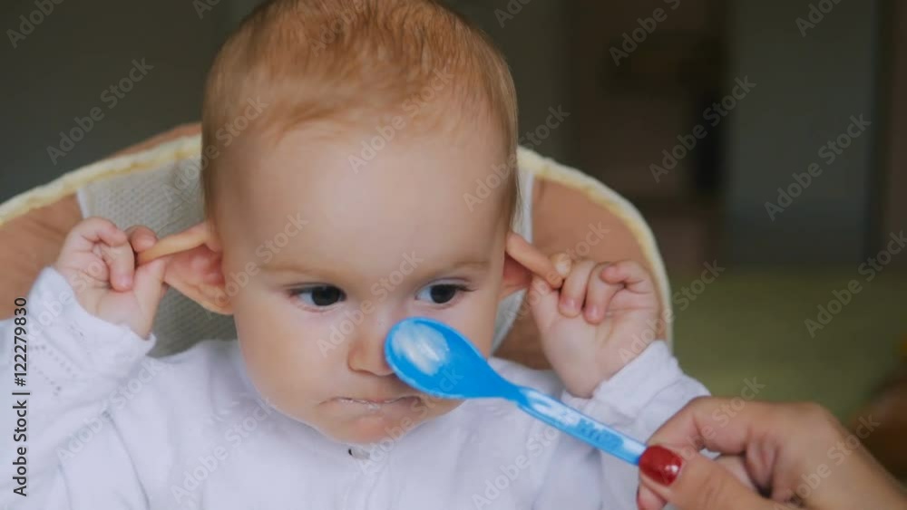 Little girl eating yogurt in Child seat and smiling. Happy family concept. Mother with spoon and baby food. Little girl eating yogurt in Child seat and smiling. Happy family concept. Mother with spoon and baby food.