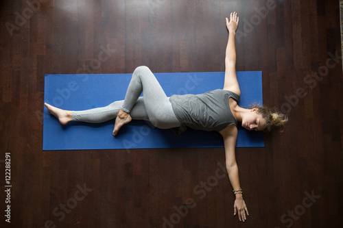 Attractive young woman working out indoors, doing yoga exercise on wooden floor, lying in Reclining Spinal Twist, Jathara Parivartanasana, resting after practice, full length, top view