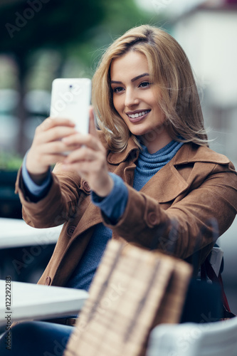 Beautiful young blonde woman siting in cafe bar, smiling and taking selfie with her cell phone.