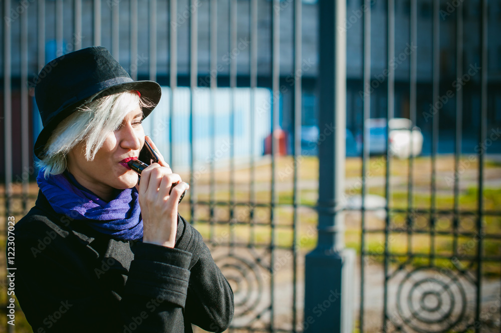 Fototapeta premium portrait of young beautiful woman with white hair, in a black coat, a skirt and a black hat, smoking an electronic cigarette, blowing the smoke vapor