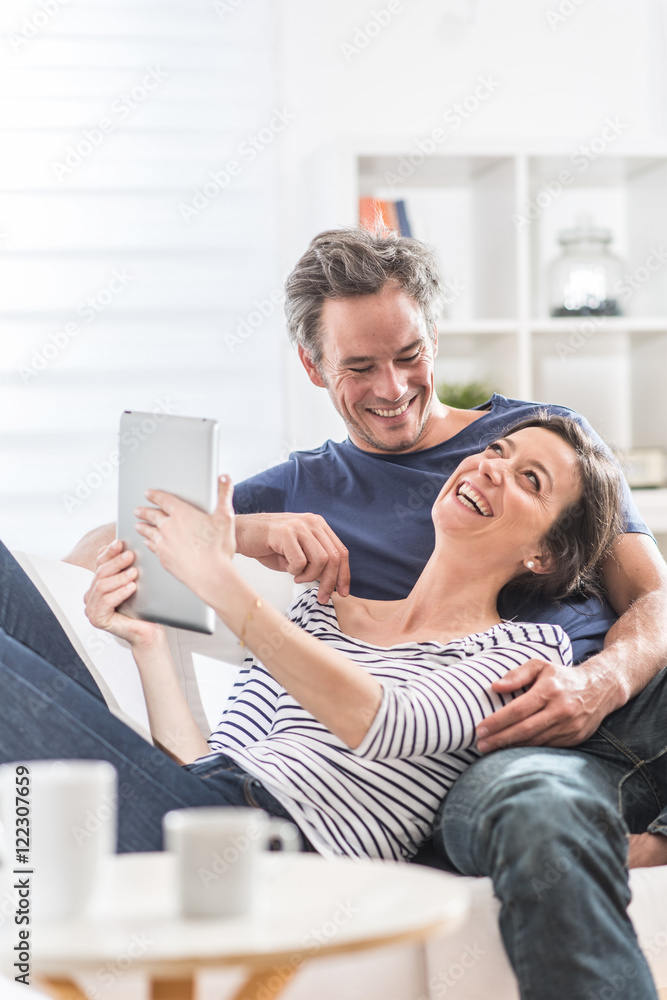 Cheerful couple sitting on a white couch at home using a tablet Cheerful couple sitting on a white couch at home using a tablet