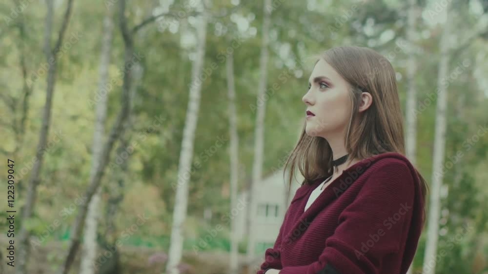 Portrait of a young model in the forest. Maroon sweater and bright lips. Girl with chokers around the neck and bright make-up on a background of trees. She looks up. Model with severe eye near trees Portrait of a young model in the forest. Maroon sweater and bright lips. Girl with chokers around the neck and bright make-up on a background of trees. She looks up. Model with severe eye near trees