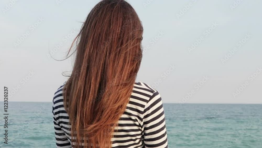 Young woman with long hair in a striped shirt looking at the sea, view from the back, slow motion Young woman with long hair in a striped shirt looking at the sea, view from the back, slow motion