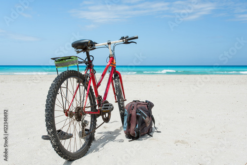 Bicycle is perked on the coast of Caribbean Sea with snow-white sand. Backpack and slates lie nearby.