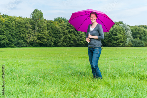 Young woman with brown hair standing on a meadow, smiling, holding a purple umbrella to protect herself from the sun
