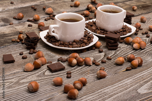 two small white cups of coffee with cocoa beans, slices of chocolate and hazelnuts on wooden background