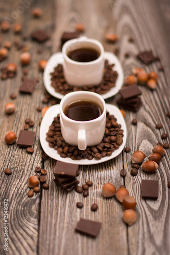 two small white cups of coffee with cocoa beans, slices of chocolate and hazelnuts on wooden background