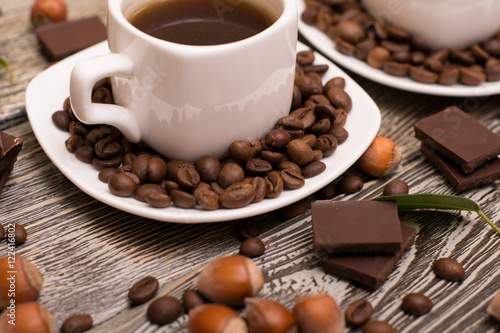 two small white cups of coffee with cocoa beans, slices of chocolate, hazelnuts and green leaves on wooden background