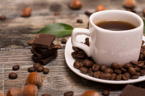 two small white cups of coffee with cocoa beans, slices of chocolate, hazelnuts and green leaves on wooden background