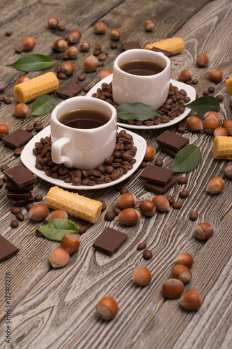 two small white cups of coffee with  cookies  cocoa beans  slices of chocolate  hazelnuts and green leaves on wooden background