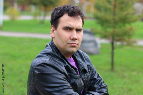 Handsome serious man in leather jacket looks at camera in park a