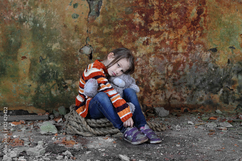 Little girl sits in basement with teddy bear in hands - orphan, homelessness, poverty, despair concept