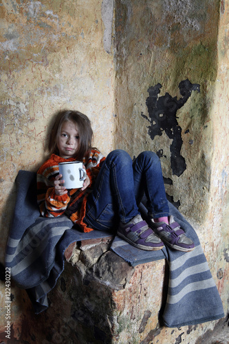 Sad little girl with dirty face sits on old blanket in basement with iron mug in hands - refugee orphan, homelessness, poverty concept
