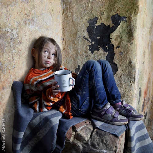 Sad little girl with dirty face sits on old blanket in basement with iron mug in hands - refugee orphan, poverty concept