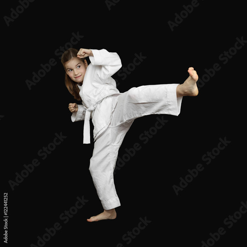 Karate practicing - little girl in sport white kimono with brave face beats foot on black background