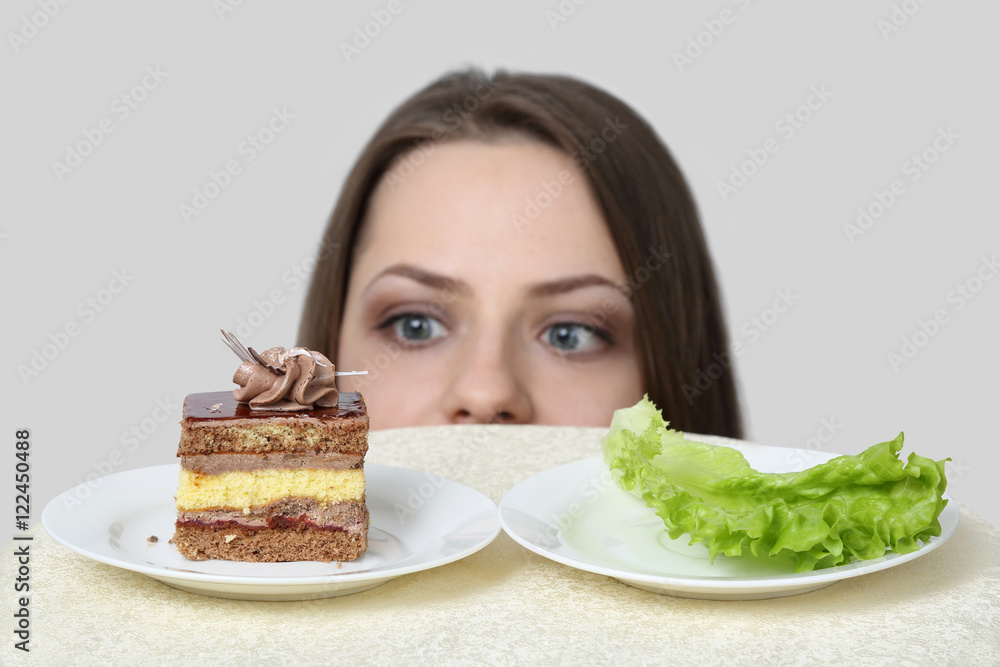 Diet, nutrition, weight loss and choice concept - Young woman chooses between cake and lettuce on gray background with focus on food Diet, nutrition, weight loss and choice concept - Young woman chooses between cake and lettuce on gray background with focus on food