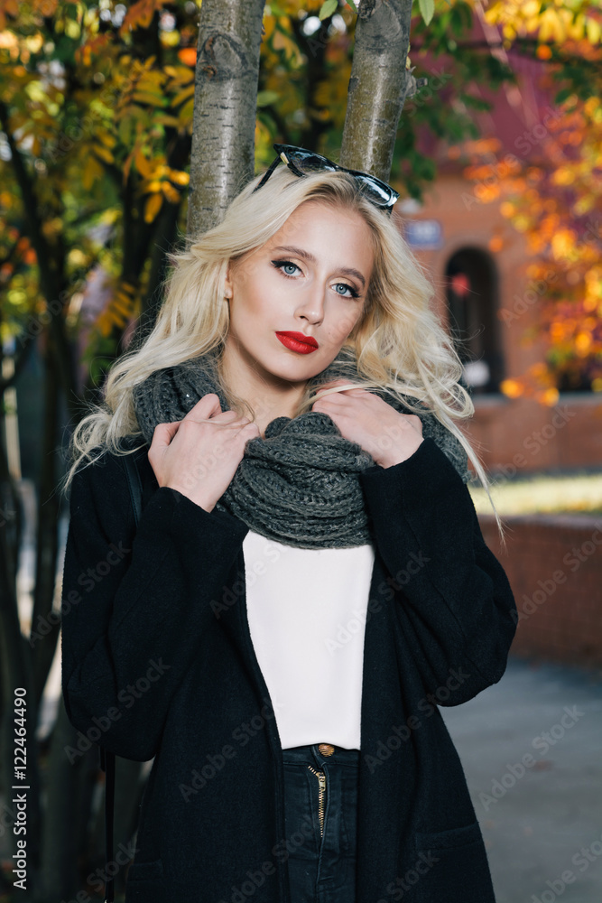 Woman with blond curly hair in park.Beautiful Portrait of sensual young model wearing a coat and grey scarf. With glamour red lips make-up, eye arrow makeup, purity skin Woman with blond curly hair in park.Beautiful Portrait of sensual young model wearing a coat and grey scarf. With glamour red lips make-up, eye arrow makeup, purity skin