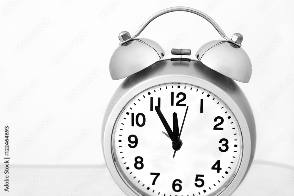Overslept all the time - slightly tilted gray alarm clock with black and white dial showing five minutes to noon. Desaturated close up view on light gray background Overslept all the time - slightly tilted gray alarm clock with black and white dial showing five minutes to noon. Desaturated close up view on light gray background