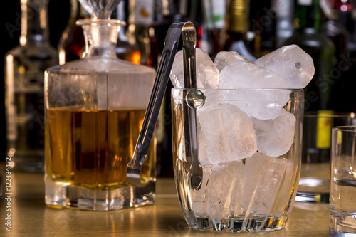 Glass ice bucket full of ice cubes in front of whiskey and other alcoholic drinks