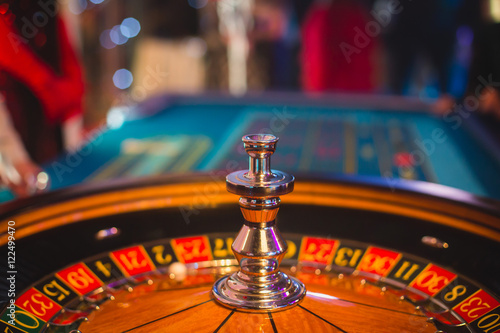 A close-up vibrant image of multicolored casino table with roulette in motion, with the hand of croupier, and a group of gambling rich wealthy people in the background 