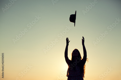 Silhouette of woman with long curly hair throwing her hat in the air at sunset outdoor.