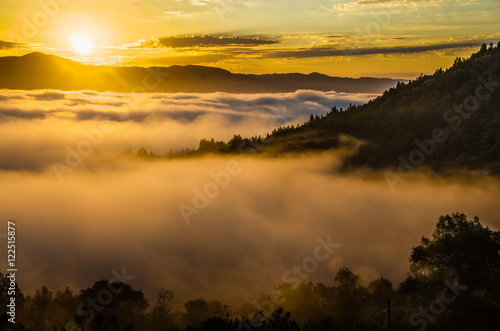 Golden sunrise shining through fog with tree covered hills in mid ground