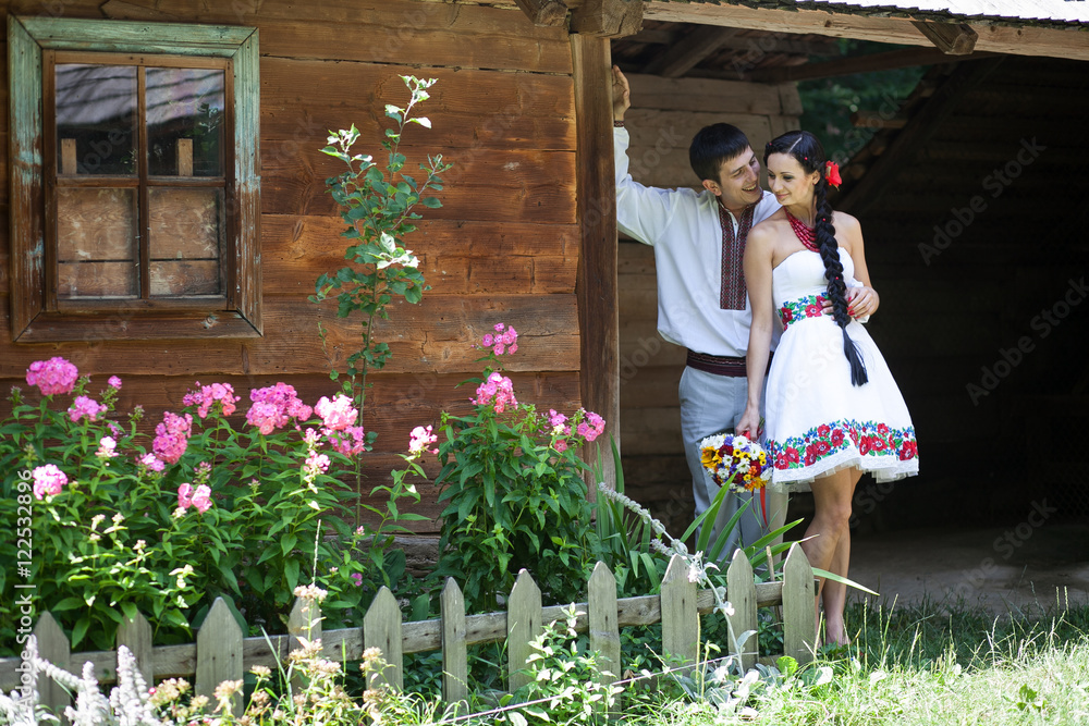 Fototapeta premium Groom hugs bride from behind while leaning to the ethnic house