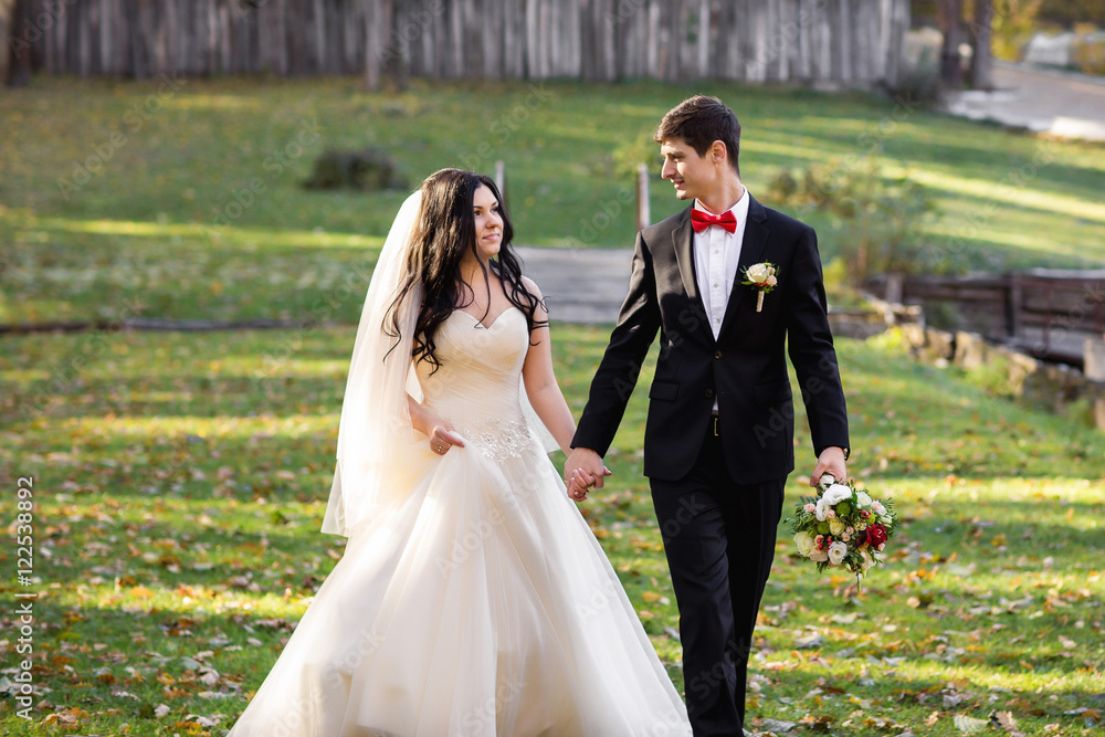 Handsome groom in black suite and red bowtie walking with happy bride in peach wedding dress in the autumn park