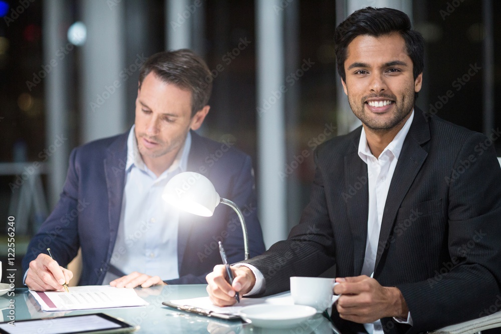 Businessman smiling at camera while colleague writing on paper Businessman smiling at camera while colleague writing on paper