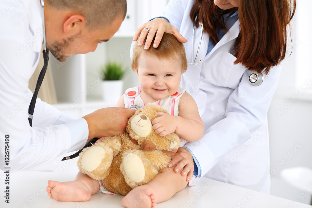 Two pediatricians are taking care of baby in hospital. Little girl is being examining by doctor with stethoscope. Health care, insurance and help concept. Two pediatricians are taking care of baby in hospital. Little girl is being examining by doctor with stethoscope. Health care, insurance and help concept.