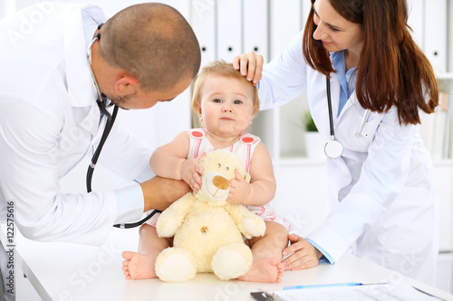 Two pediatricians are taking care of baby in hospital. Little girl is being examining by doctor with stethoscope. Health care, insurance and help concept.