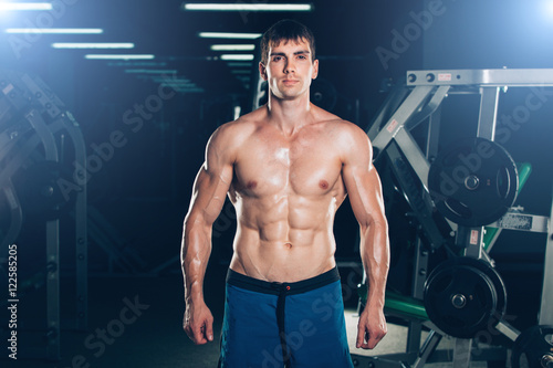 Young Man Standing Strong In The Gym And Flexing Muscles - Muscular Athletic Bodybuilder Fitness Model Posing After Exercises.