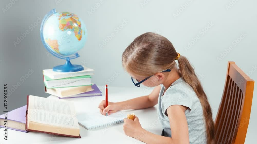School girl four-eyes writing in her notebook and eating croissant and smiling at camera. Child sitting at desk isolated on white background. Thumbs up. Ok School girl four-eyes writing in her notebook and eating croissant and smiling at camera. Child sitting at desk isolated on white background. Thumbs up. Ok