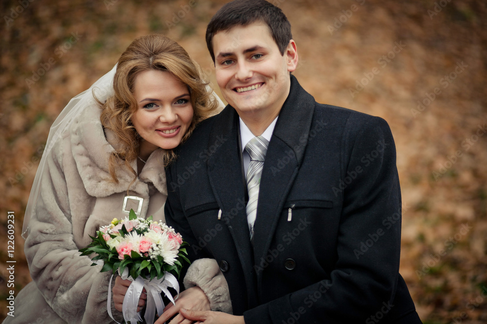 Prim newlyweds dressed in warm clothes sit in the park full of f Prim newlyweds dressed in warm clothes sit in the park full of f