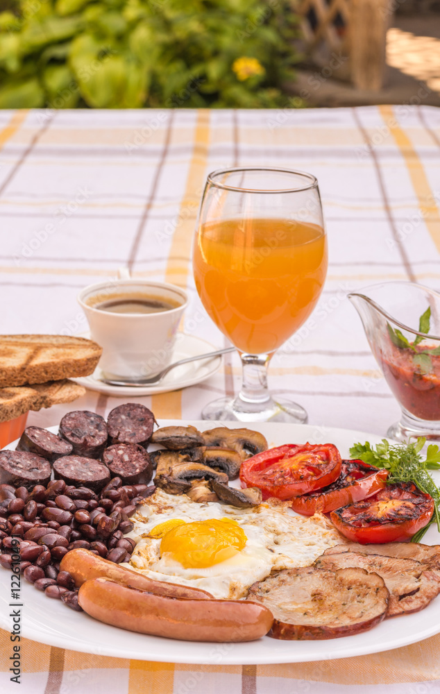 Full English fried breakfast with bacon, egg, sausages, black pudding, mushrooms, grilled tomatoes and baked beans. Closeup with toasts, cup of coffee, orange juice and home made  red sauce.